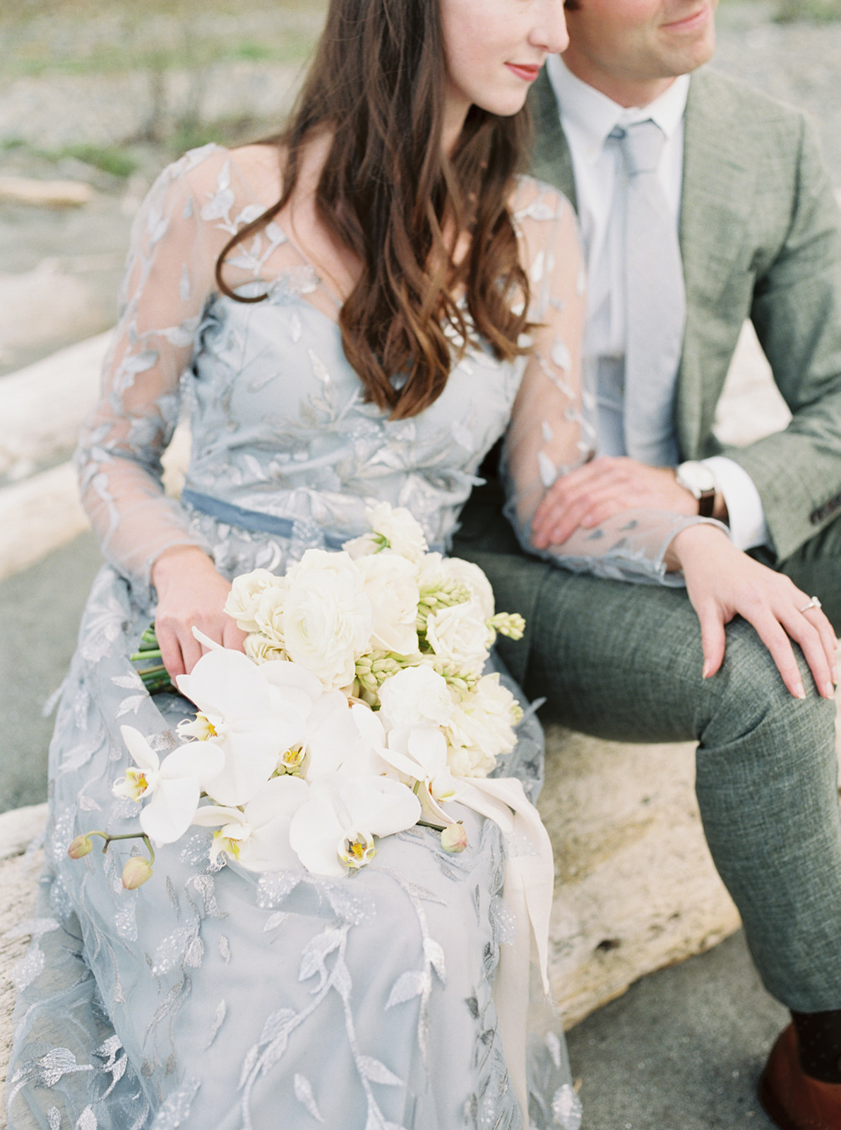 A couple sitting on a driftwood beach holindg a bouquet
