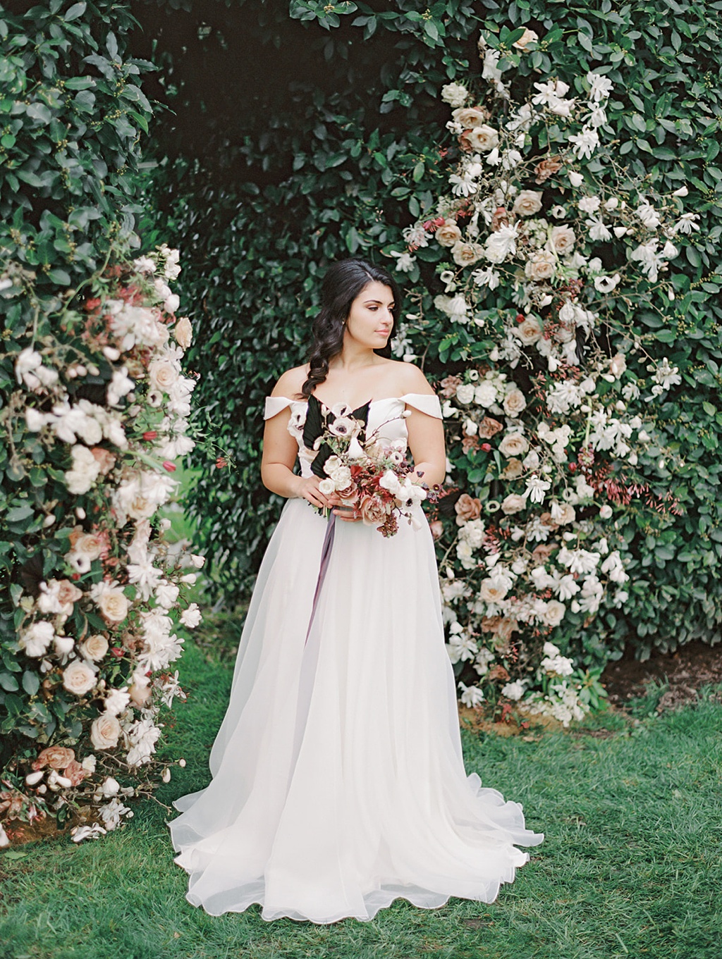 A bride holding a bouquet stands in front of a manicured hedge "doorway" to the vineyard at Chateau Lill