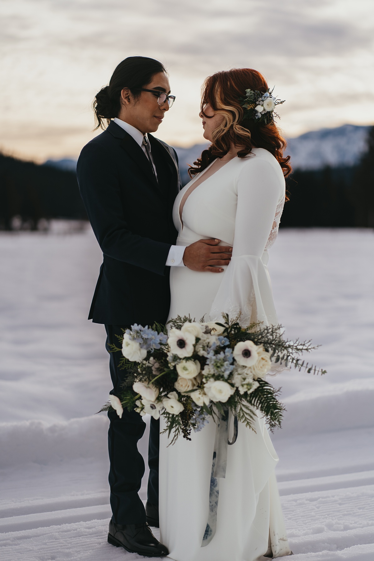 A couple embracing at sunset on their wedding day, in the snow, holding bouquet