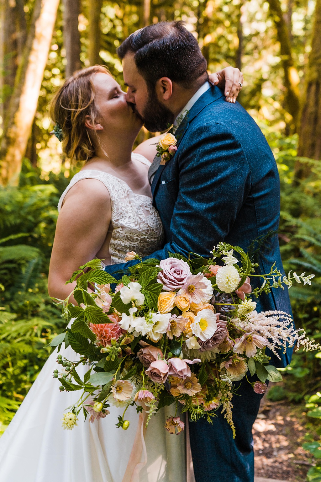 A bride and groom kiss in the woods holding a bouquet
