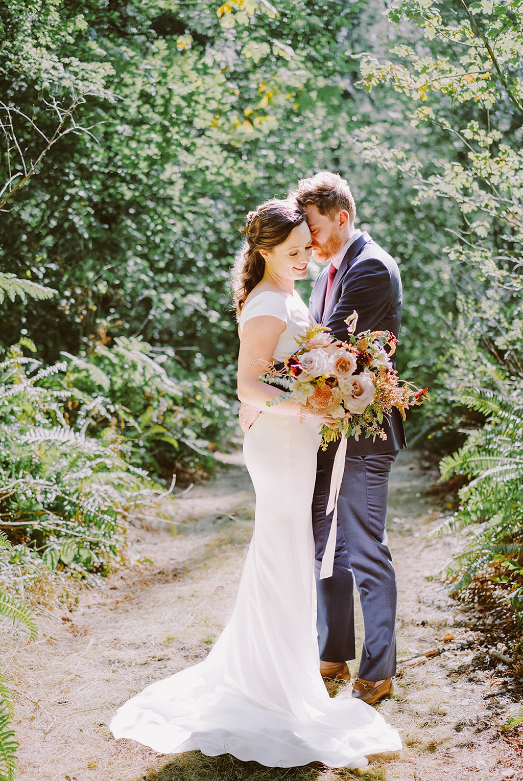 The couple stands in the woods, holding a summer wedding bouquet 