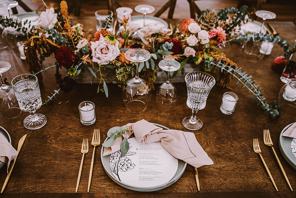 A long farm table is set with a large floral arrangement, flatware, plates, napkins, and botanical sprigs